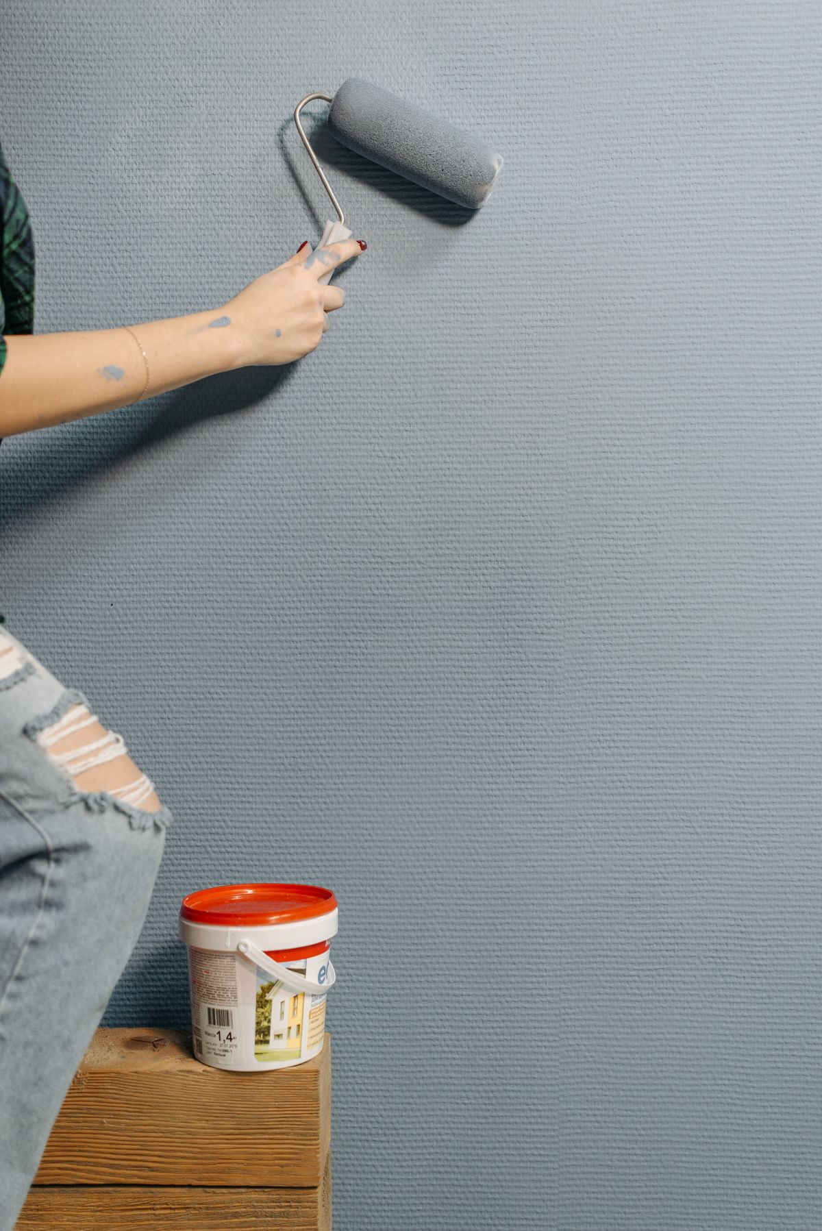 A hand rolling gray paint onto an interior wall, with a paint can below
