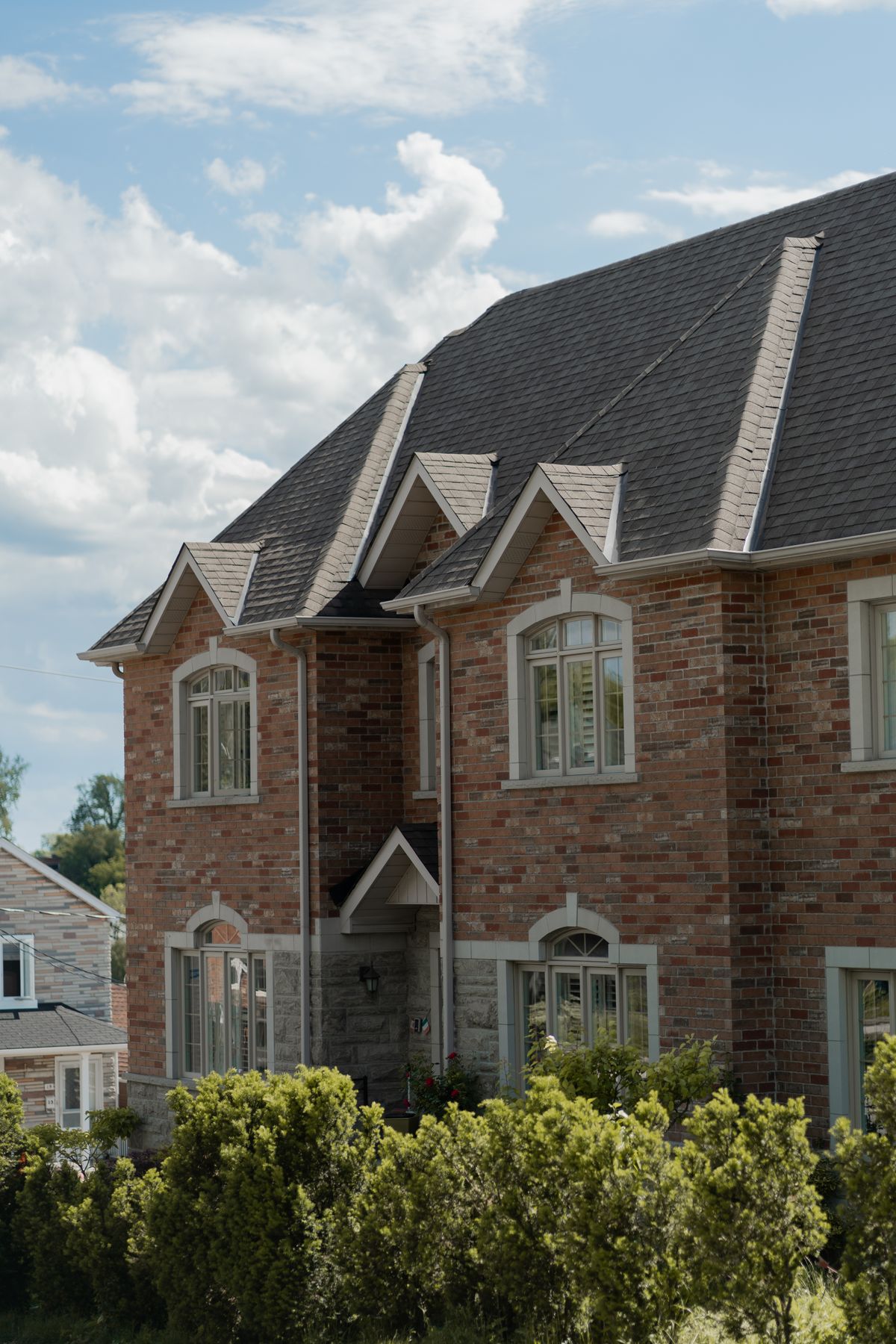 A large brick house with a steep shingled roof and green shrubs
